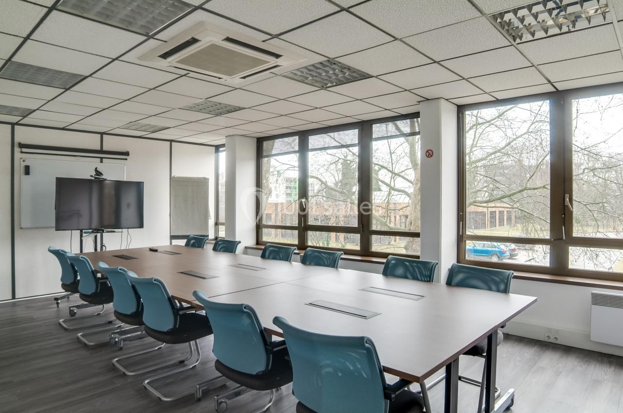 Salle de réunion lumineuse avec une grande table rectangulaire, des chaises bleues et un écran de présentation.