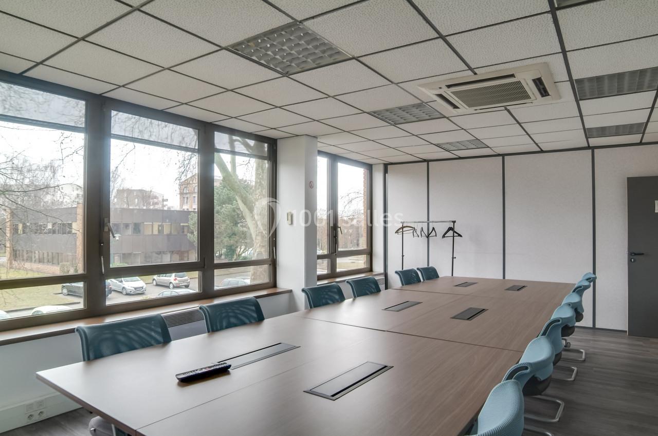 Salle de réunion lumineuse avec grande table en bois, chaises bleues, fenêtres donnant sur un extérieur arboré.