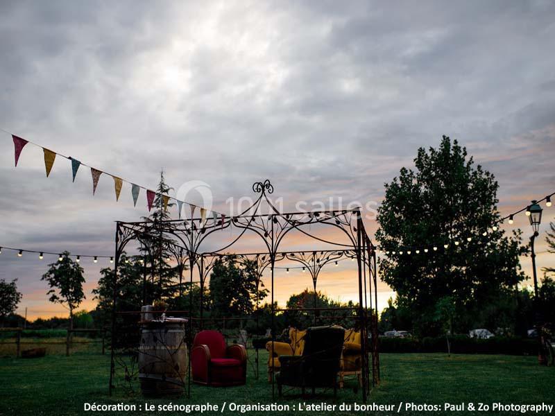 Structure métallique décorative avec fauteuils et guirlandes lumineuses, installée dans un jardin au coucher du soleil.