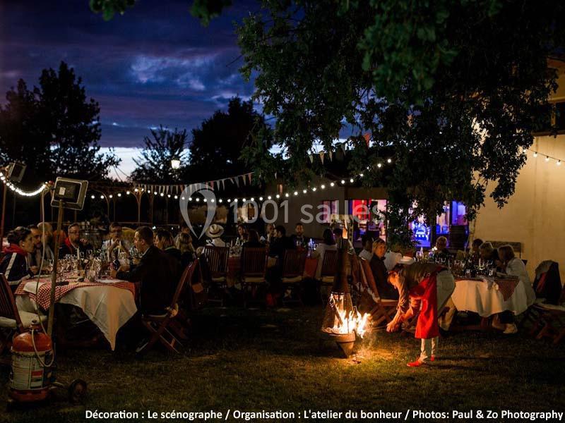 Des convives dînent en extérieur sous des guirlandes lumineuses, tandis qu'un enfant alimente un feu de camp.