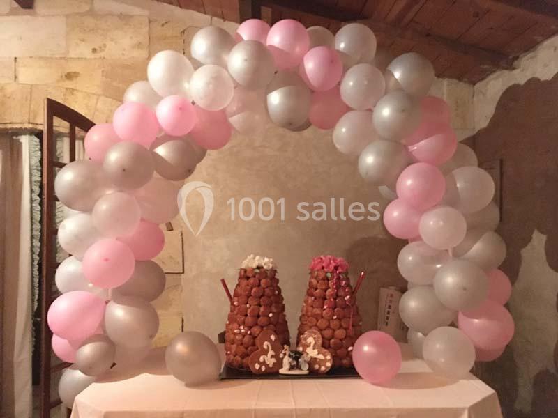 Arc de ballons roses et gris encadrant deux pièces montées en choux sur une table dans une salle en pierre.