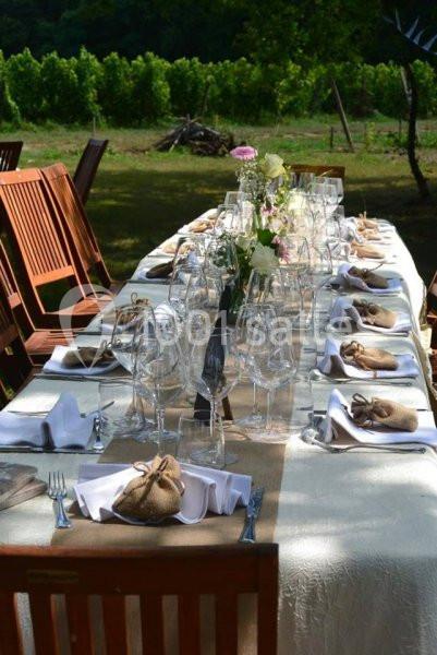 Table extérieure dressée avec nappes blanches, verres, couverts et petits sachets en toile, entourée de chaises en bois.