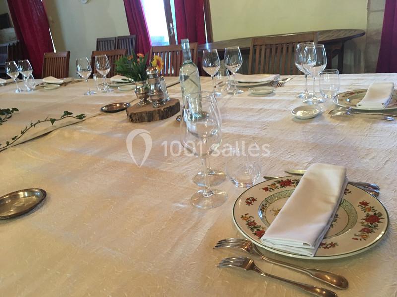 Table dressée avec nappes blanches, assiettes décorées, verres et couverts, entourée de chaises en bois.