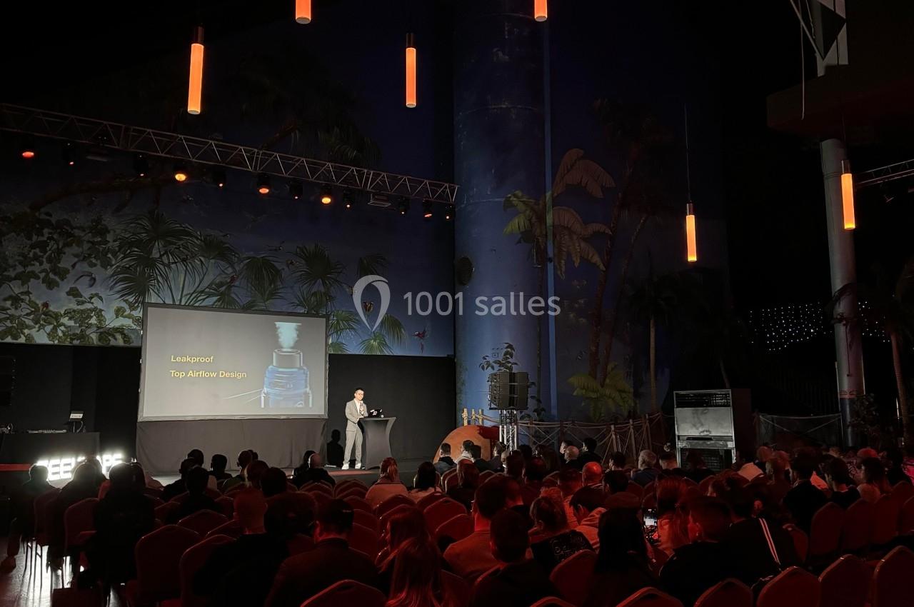 Conférence en intérieur avec un orateur sur scène, public assis et éclairage tamisé dans une grande salle.