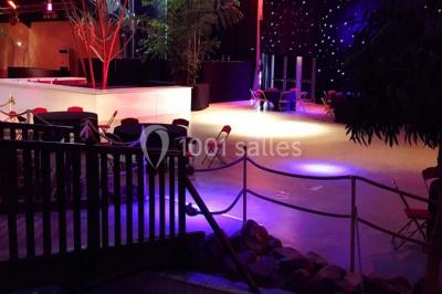 Piscine intérieure décorée de plantes tropicales, éclairée par des lumières colorées et entourée d'un pont en bois.