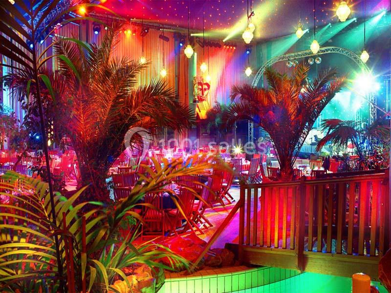 Salle décorée avec des palmiers, éclairages colorés et tables dressées pour un événement festif en intérieur.