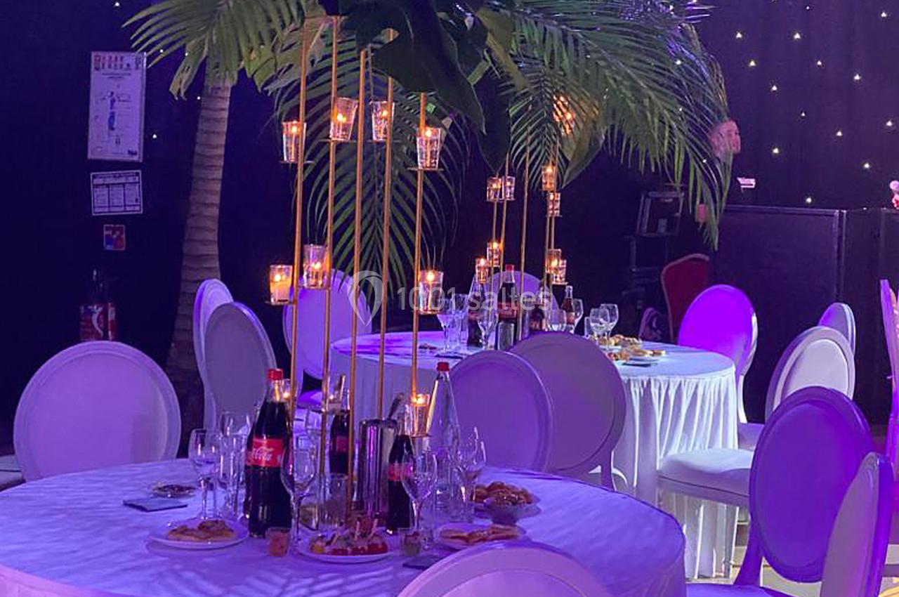 Tables rondes décorées avec des bougies suspendues, des bouteilles et des verres, sous des palmiers artificiels dans une…