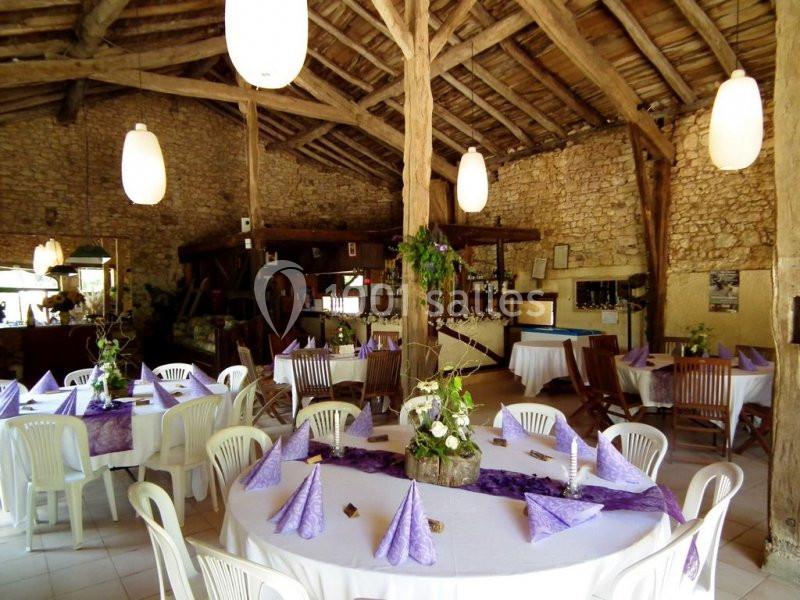 Salle rustique avec murs en pierre, poutres apparentes et tables rondes décorées de nappes blanches et violettes.