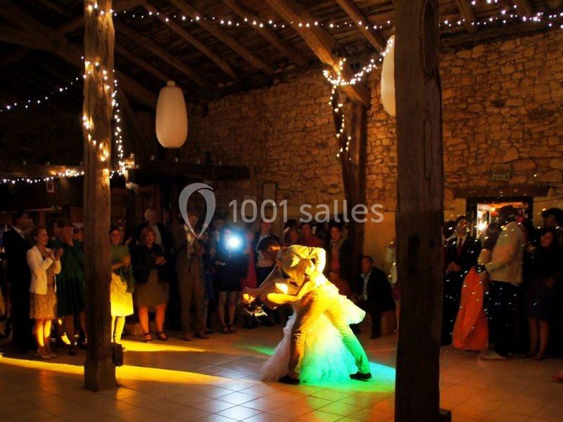 Un couple de mariés danse au centre d'une salle rustique éclairée par des guirlandes lumineuses, entouré d'invités.