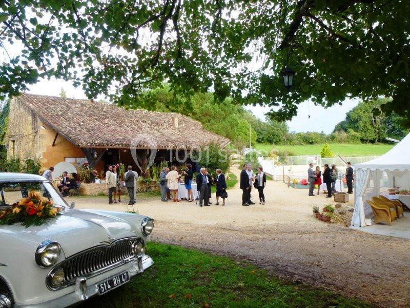 Personnes rassemblées devant une maison en pierre et une tente blanche dans un cadre champêtre, avec une voiture ancienne au…