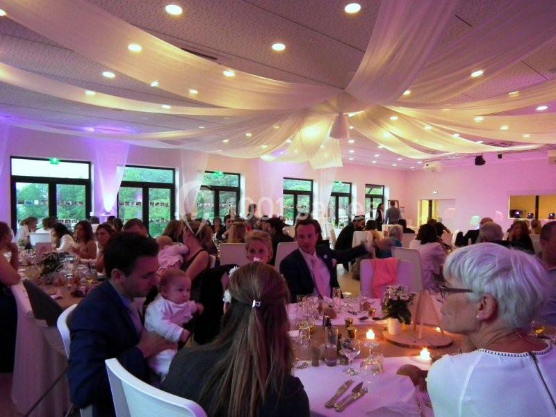 Salle de réception décorée avec des drapés blancs, éclairage tamisé, invités assis à des tables lors d'un événement.