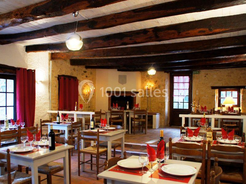Salle de restaurant rustique avec poutres apparentes, tables dressées avec nappes rouges et lumière chaleureuse.