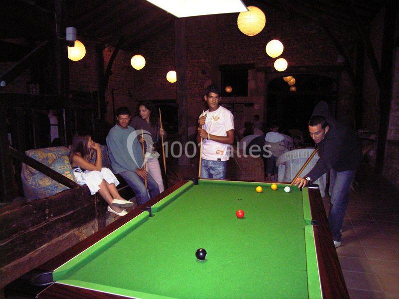 Des personnes jouent au billard dans une pièce éclairée par des lampes suspendues, avec des spectateurs assis.