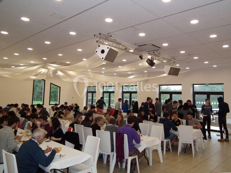 Des personnes assises et debout dans une grande salle lumineuse, partageant un repas autour de tables blanches.