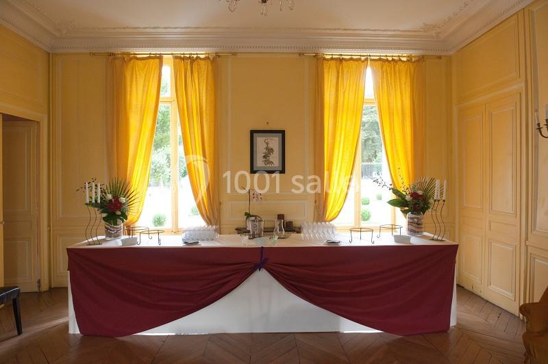 Table décorée avec une nappe bordeaux, ornée de fleurs et de chandeliers, devant des fenêtres aux rideaux jaunes.