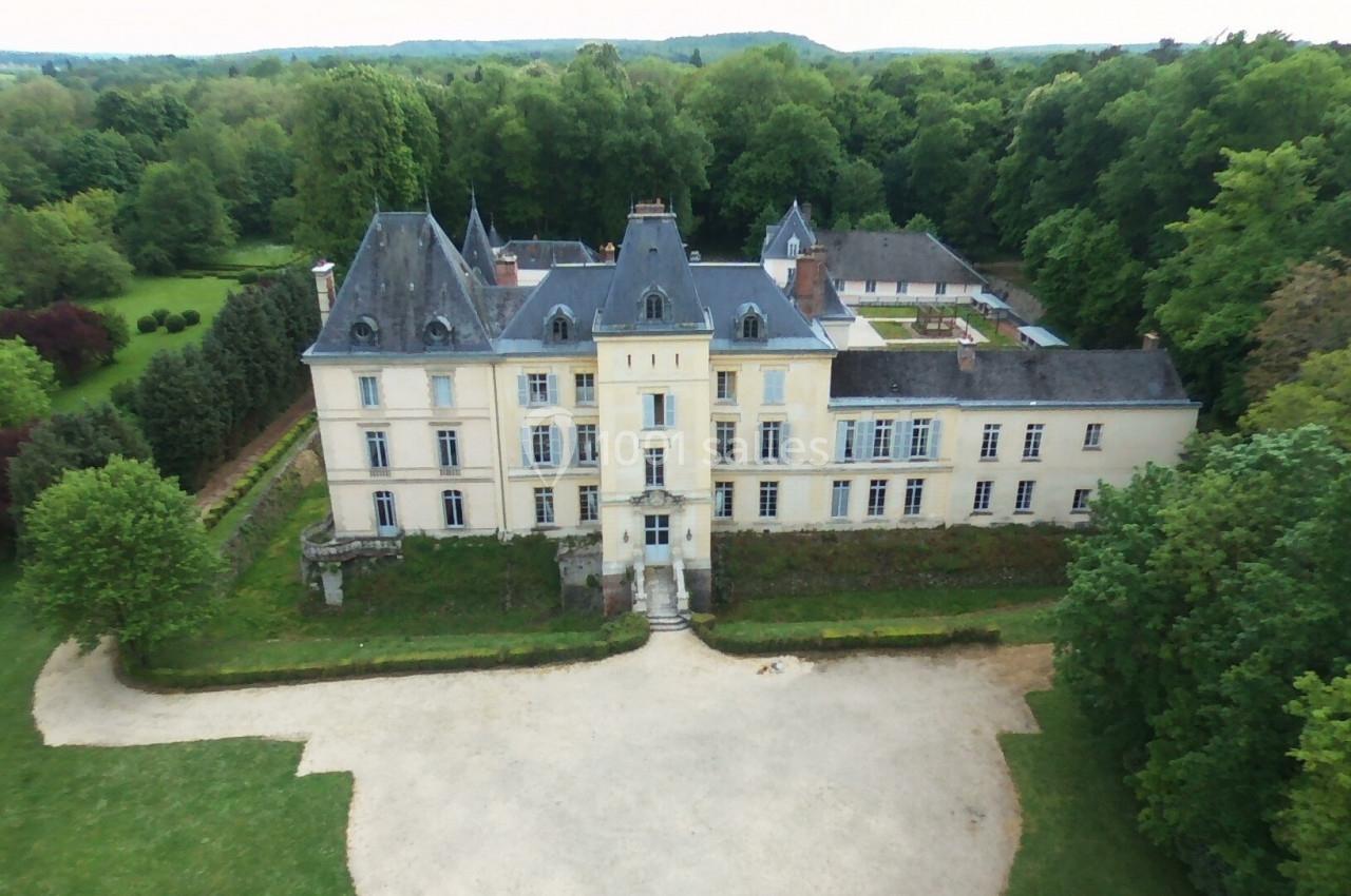 Vue aérienne d'un château entouré de jardins verdoyants et d'une forêt, avec une cour dégagée à l'avant.