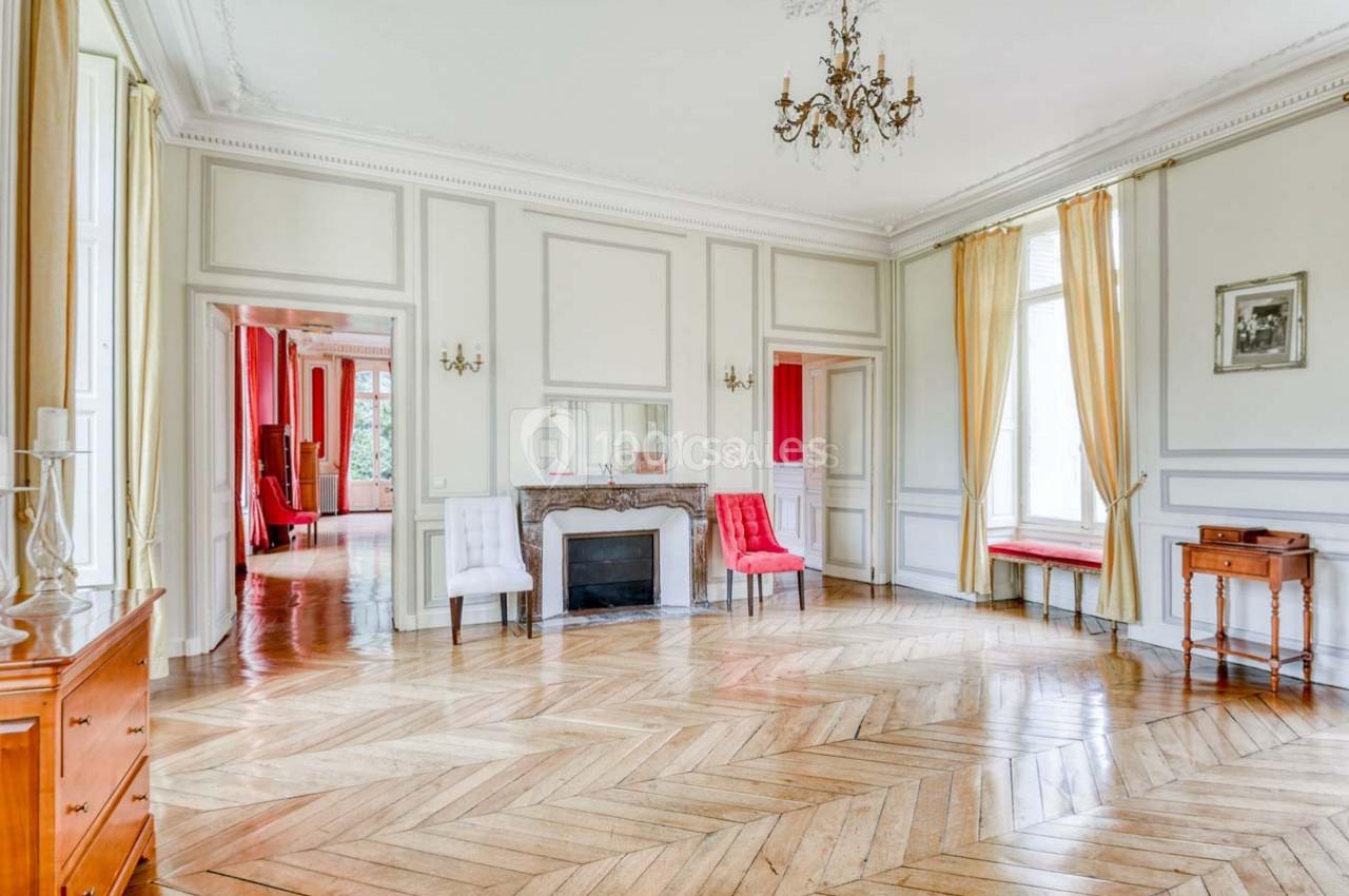 Salon lumineux avec parquet en bois, cheminée décorative, chaises et grandes fenêtres encadrées de rideaux jaunes.