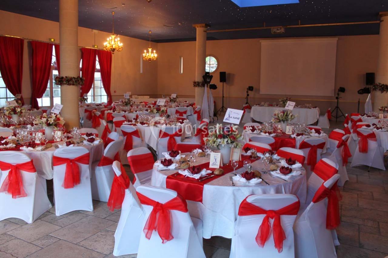 Salle de réception décorée pour un événement, avec des tables rondes habillées de nappes blanches et nœuds rouges.
