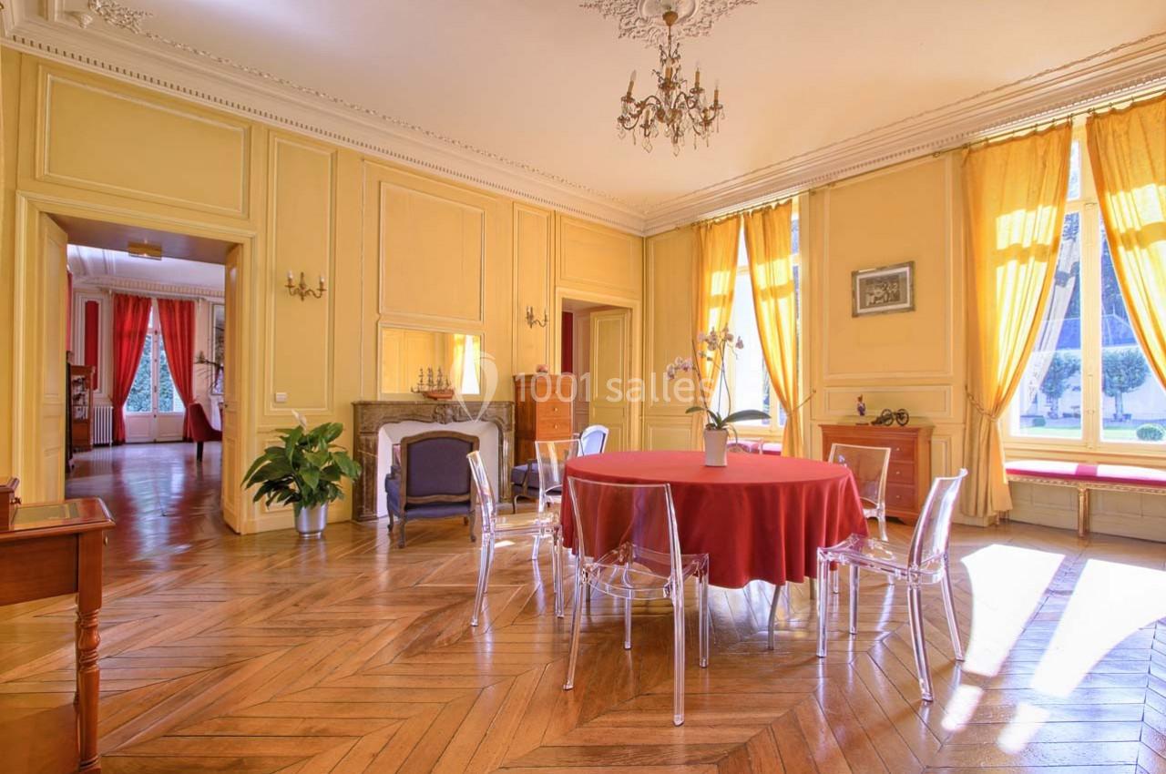 Salle lumineuse avec parquet en bois, murs jaunes, table ronde avec nappe rouge et chaises transparentes.