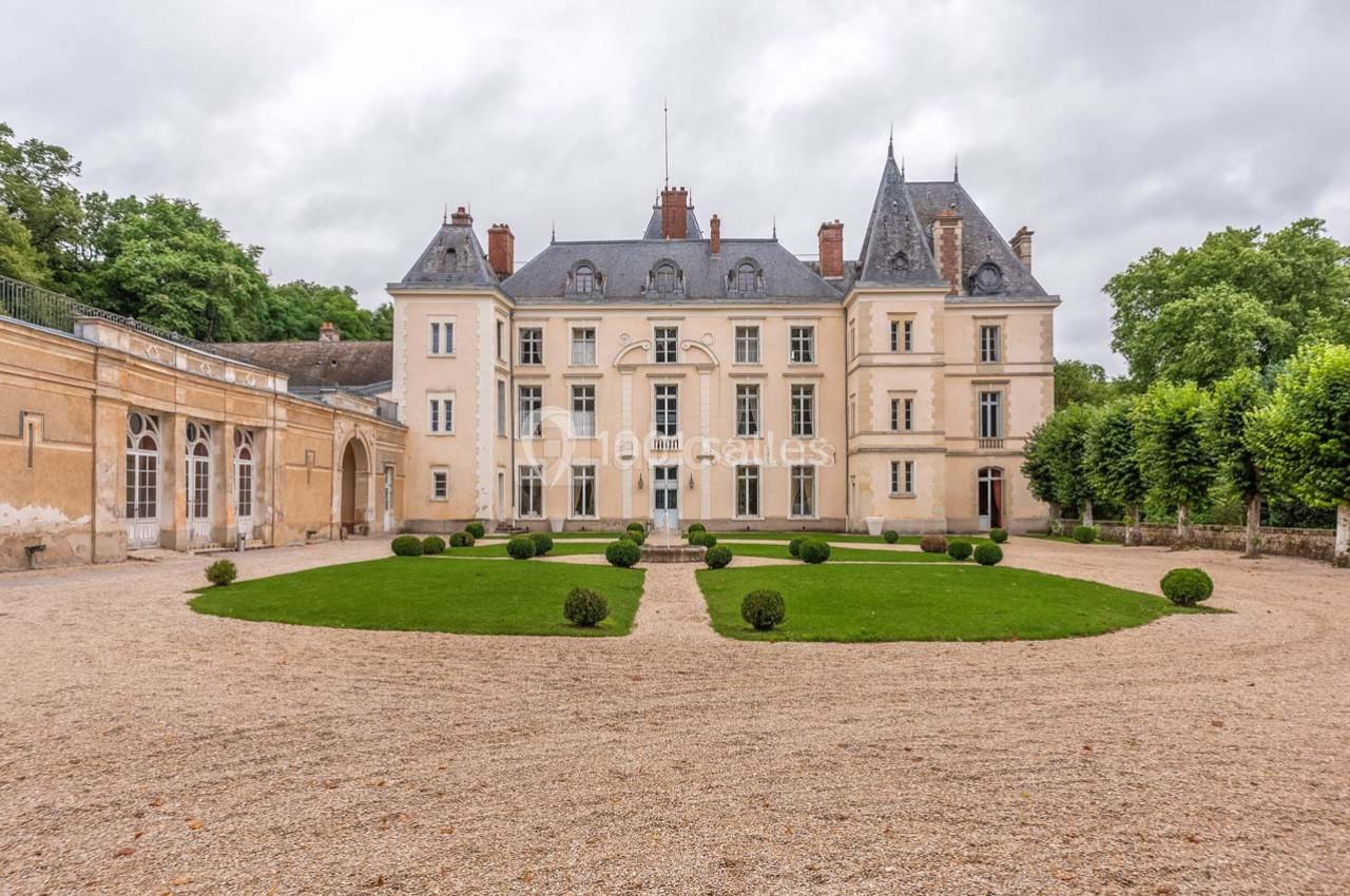 Façade d'un château historique entouré d'une cour gravillonnée et d'un jardin avec pelouse et arbustes taillés.