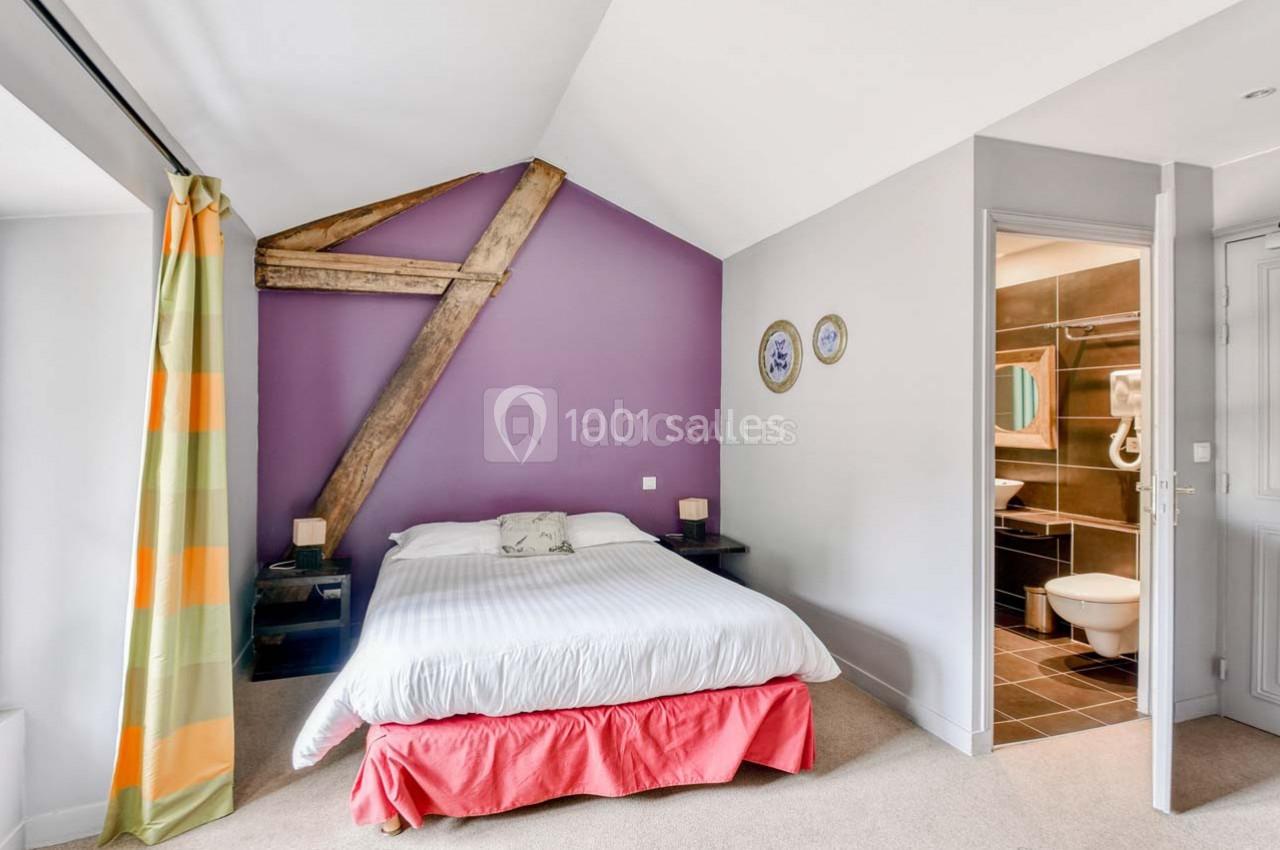 Chambre lumineuse avec lit double, mur violet avec poutre apparente, et salle de bain attenante visible à droite.