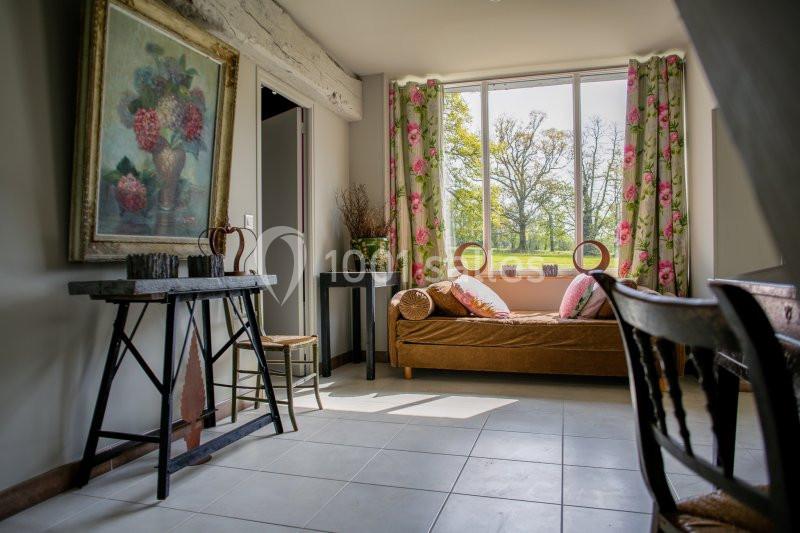 Salon lumineux avec canapé, table en bois, tableau floral et grande fenêtre donnant sur un jardin verdoyant.