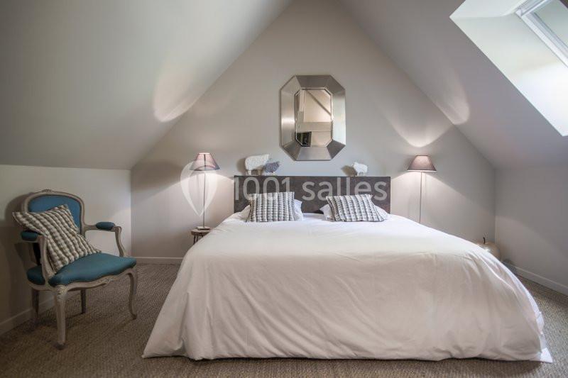 Chambre mansardée avec lit double, coussins rayés, fauteuil bleu et lumière naturelle entrant par une fenêtre de toit.