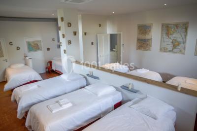 Dortoir lumineux avec plusieurs lits simples alignés, draps blancs et cartes géographiques accrochées aux murs.