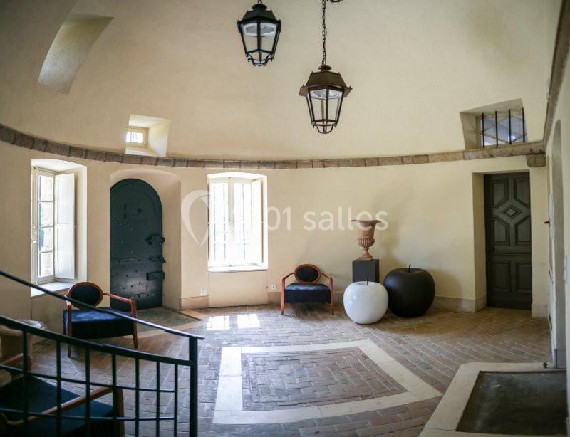 Salle lumineuse avec murs clairs, sol en briques, deux fenêtres, des chaises, des objets décoratifs et des portes.
