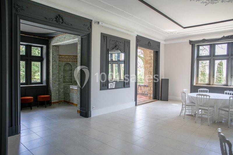 Salle lumineuse avec grandes fenêtres, murs clairs, moulures décoratives, et mobilier blanc dans un espace élégant.