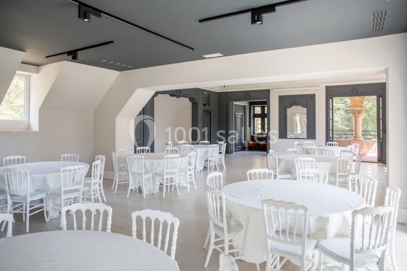 Salle lumineuse avec tables rondes dressées de nappes blanches et chaises blanches, grandes fenêtres en arrière-plan.