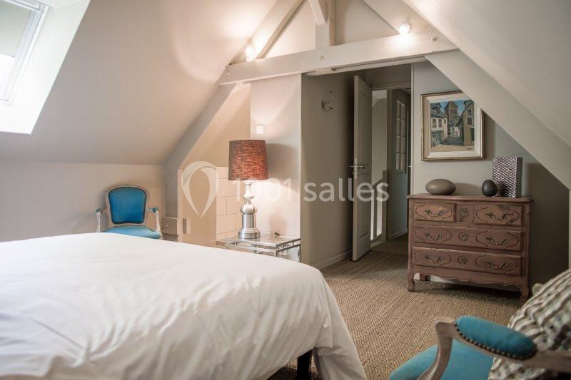Chambre mansardée avec lit blanc, fauteuils bleus, commode en bois et lampe sur table en verre.