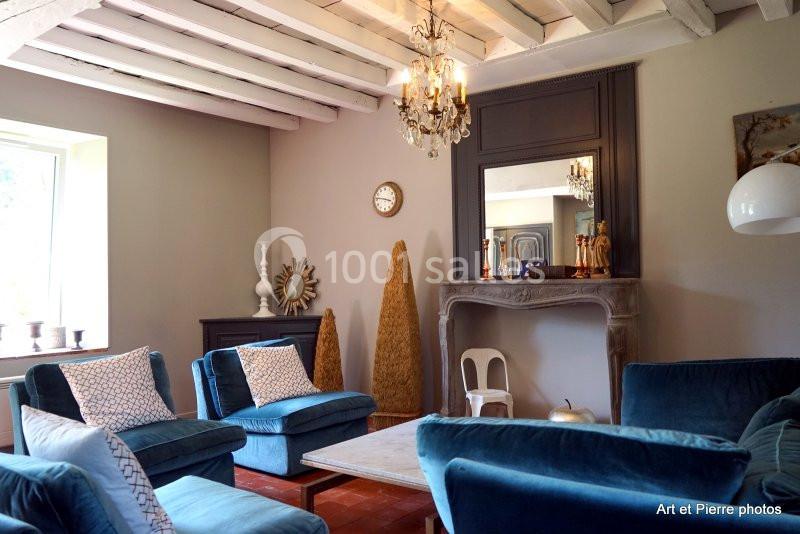 Salon lumineux avec poutres apparentes, fauteuils en velours bleu, cheminée en pierre et décoration élégante.