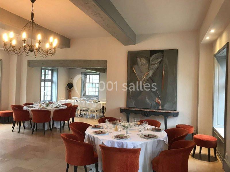 Salle de réception lumineuse avec tables rondes dressées, chaises orange et un grand tableau moderne au mur.