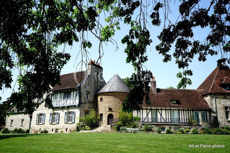 Façade d'un bâtiment ancien en pierre avec colombages, toit en tuiles et jardin verdoyant au premier plan.
