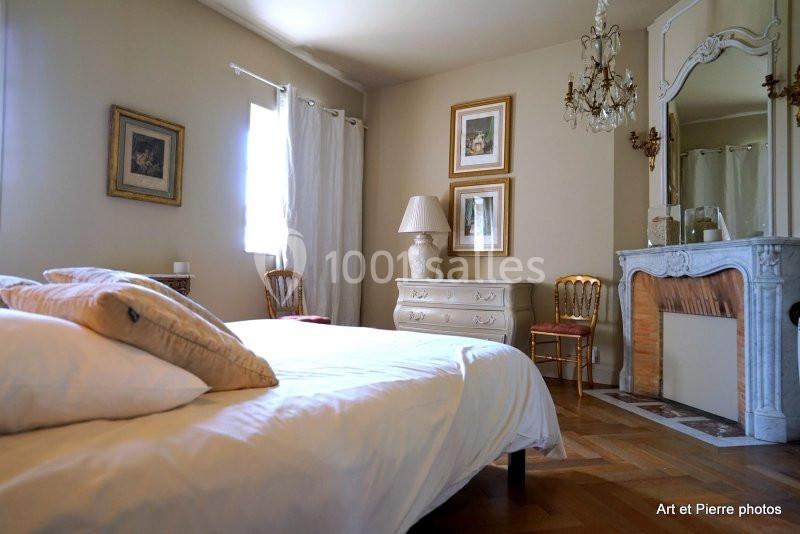 Chambre lumineuse avec lit double, cheminée décorative, commode blanche et cadres accrochés sur des murs beiges.