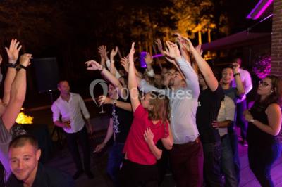Vue nocturne d'une maison en bois éclairée, entourée d'arbres illuminés, sous un ciel nuageux avec la lune visible.
