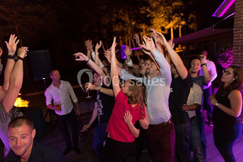 Groupe de personnes dansant et levant les bras lors d'une soirée en extérieur, éclairée par des lumières colorées.