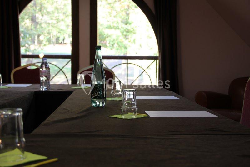 Salle de réunion avec une table en U, des verres, des feuilles blanches et une bouteille d'eau au centre.
