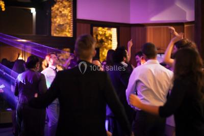 Vue nocturne d'une maison en bois éclairée, entourée d'arbres illuminés, sous un ciel nuageux avec la lune visible.