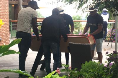 Vue nocturne d'une maison en bois éclairée, entourée d'arbres illuminés, sous un ciel nuageux avec la lune visible.