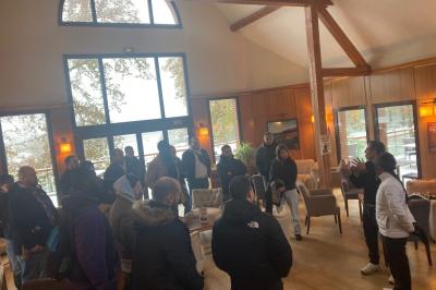 Vue nocturne d'une maison en bois éclairée, entourée d'arbres illuminés, sous un ciel nuageux avec la lune visible.