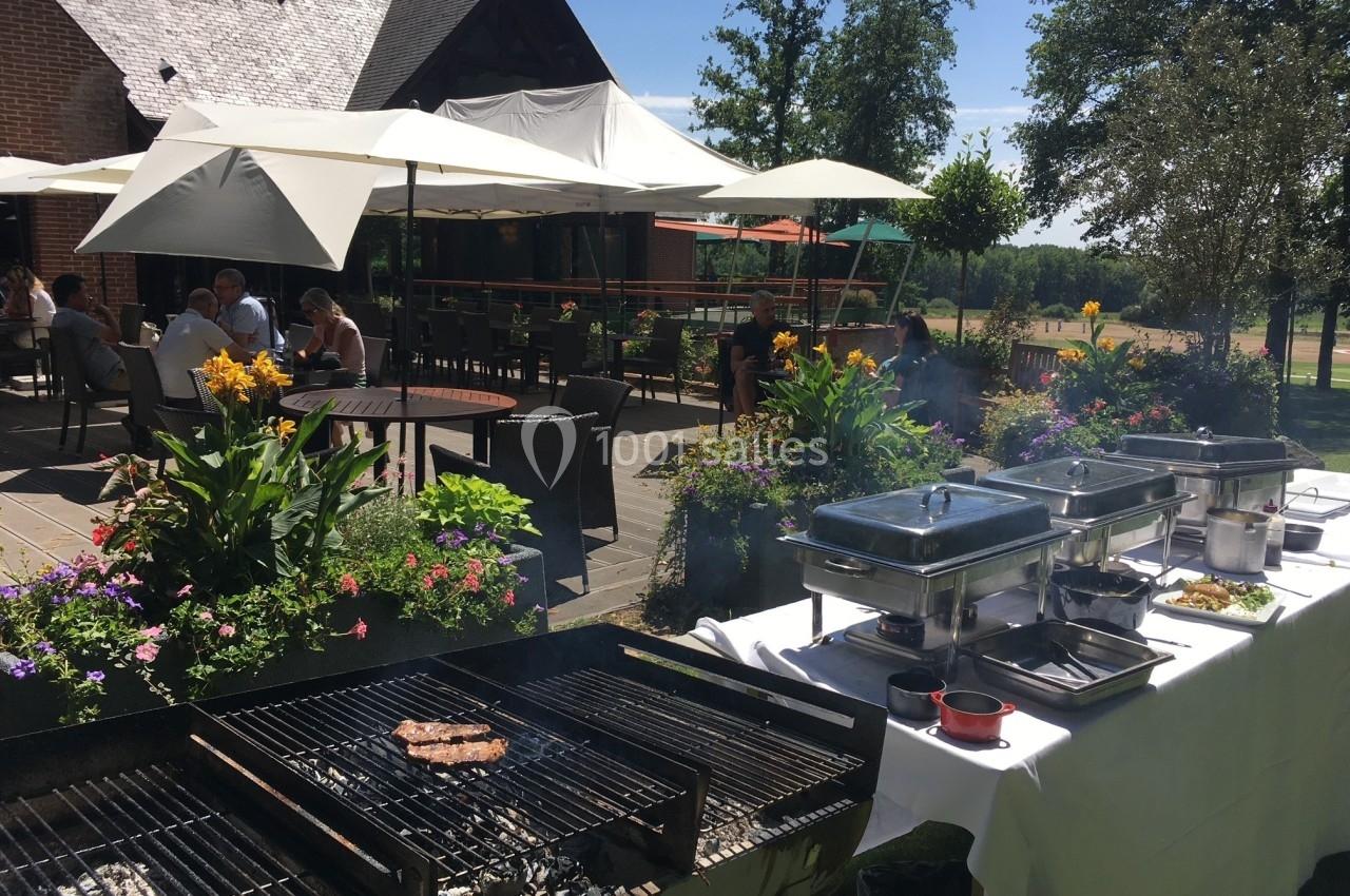 Barbecue en plein air sur une terrasse fleurie avec des tables ombragées et des convives en arrière-plan.