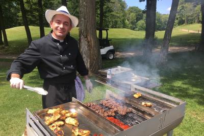 Vue nocturne d'une maison en bois éclairée, entourée d'arbres illuminés, sous un ciel nuageux avec la lune visible.