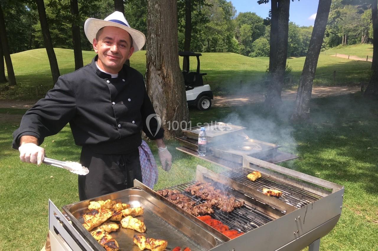 Un cuisinier en tenue noire prépare des grillades variées sur un barbecue en plein air, près d'un terrain verdoyant.