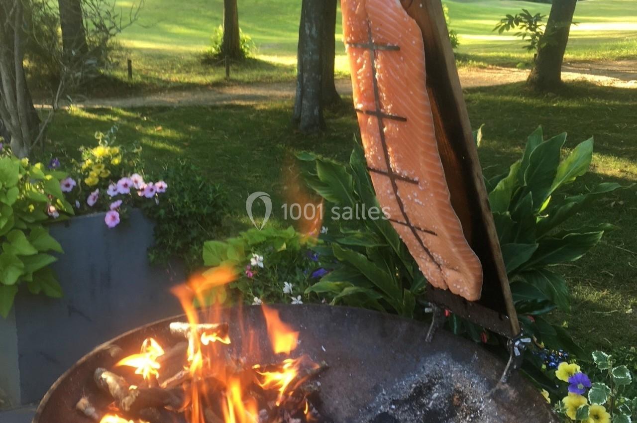 Saumon en cuisson verticale sur une planche de bois près d'un feu de bois, dans un jardin verdoyant.