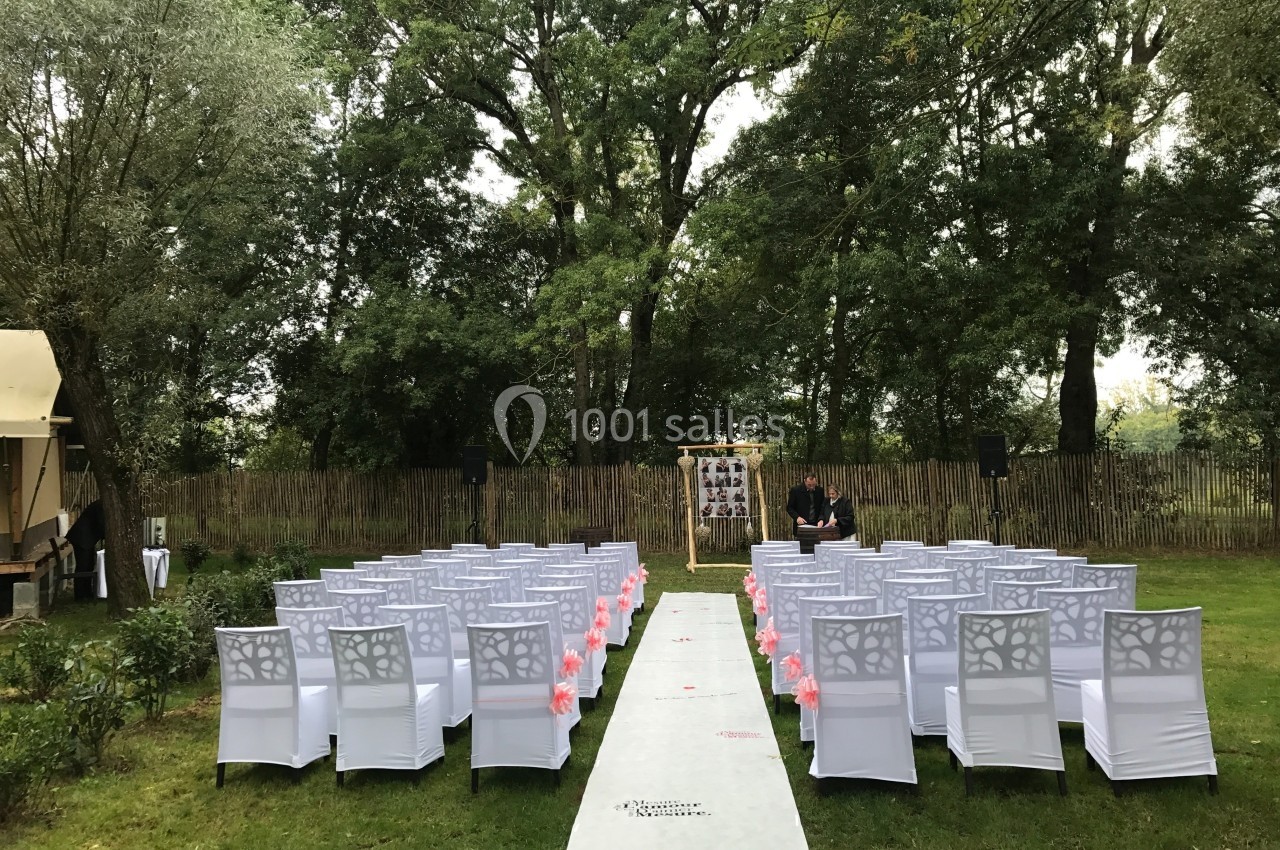 Allée centrale bordée de chaises blanches décorées, menant à une arche dans un jardin verdoyant.