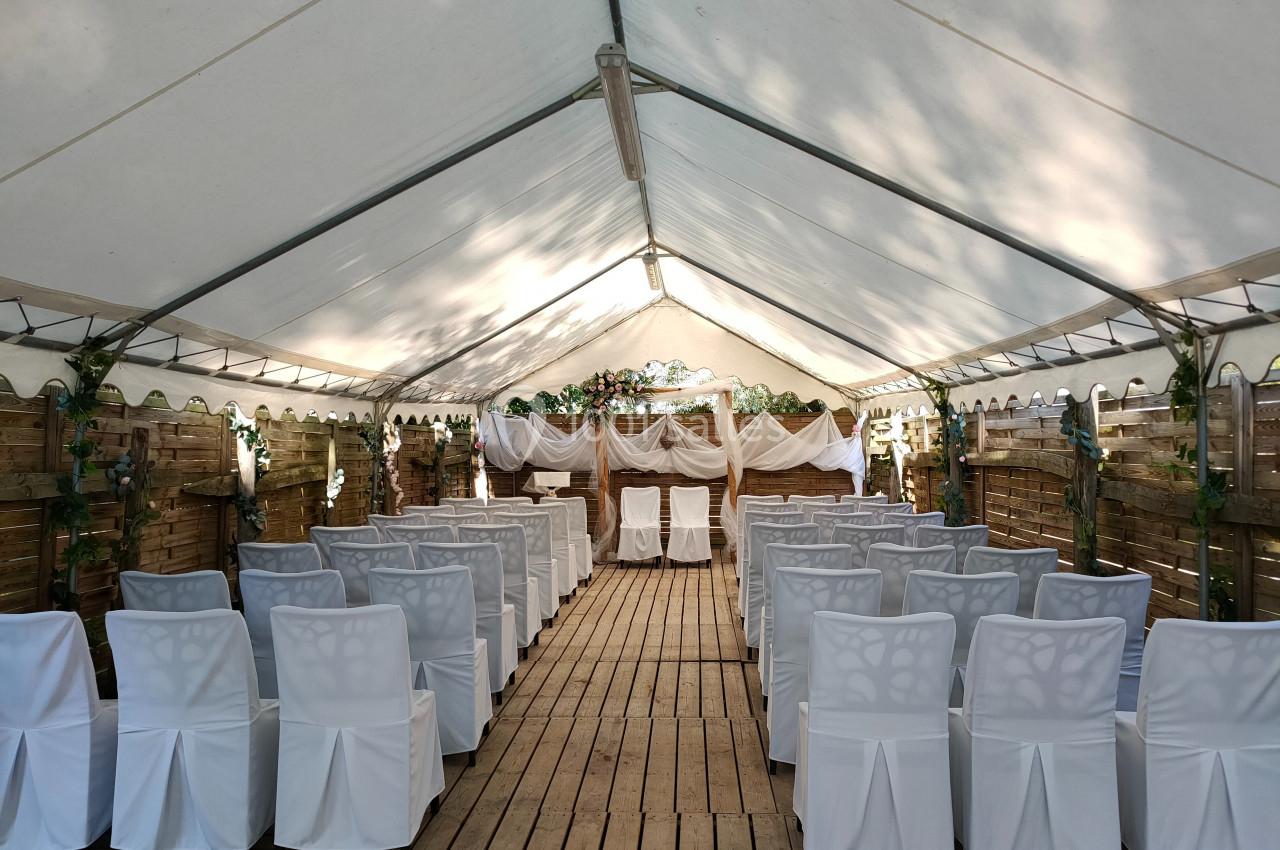 Salle de cérémonie en plein air sous une tente, avec des chaises blanches alignées et une décoration en bois et tissu.