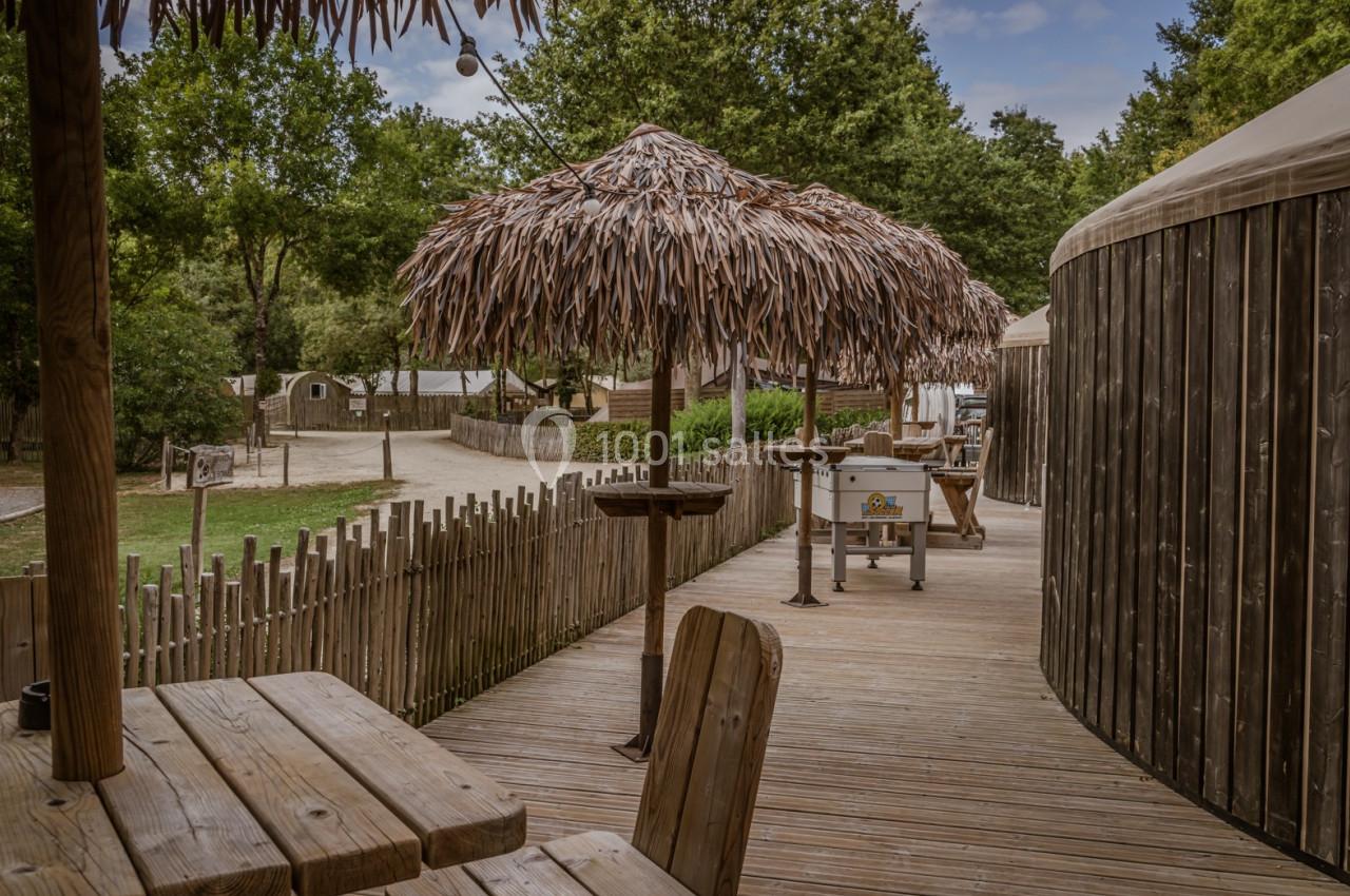 Terrasse en bois avec tables, chaises, parasols en feuilles et chemin bordé de cabanes dans un cadre boisé.
