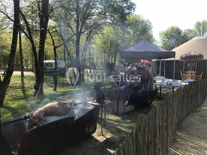 Des cochons rôtissent sur des broches au-dessus de braises dans un espace extérieur boisé avec des stands et une tente.
